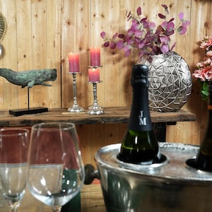 May include: A silver metal ice bucket with two bottles of champagne inside. The ice bucket is on a wooden table with a silver metal whale sculpture, candlesticks, and a silver metal vase with purple flowers.
