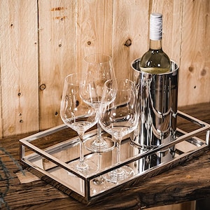 Peut inclure: Un ensemble de présentation de vin sur une surface en bois. L'ensemble comprend un seau à vin argenté avec une bouteille de vin à l'intérieur, trois verres à vin et un plateau miroir avec un cadre argenté. En arrière-plan, un mur en bois.