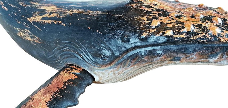 May include: Close-up of a wooden whale sculpture with a distressed black and copper paint finish. The whale's eye and mouth are carved into the wood, and the tail fin is partially visible.