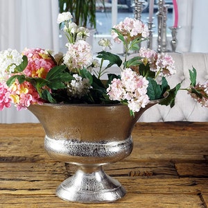 Puede incluir: Un jarrón de metal plateado con una abertura ancha y redonda y una base de pedestal. El jarrón está lleno de flores rosas, blancas y amarillas.