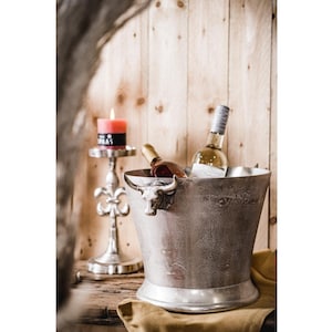 May include: A silver metal ice bucket with a bull head handle, filled with ice and two bottles of wine. A silver candle holder with a pink candle is in the background.
