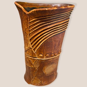 Puede incluir: Un jarrón de cerámica marrón con un diseño texturizado y abstracto. El jarrón tiene una boca ancha y una base estrecha.