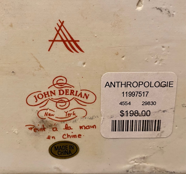 May include: A white ceramic box with a red and black label that reads "John Derian New York" and "Peint a la main en Chine." The label also has a red logo with the letter "A." The box has a price tag that reads "Anthropologie 11997517 4554 29830 $198.00".
