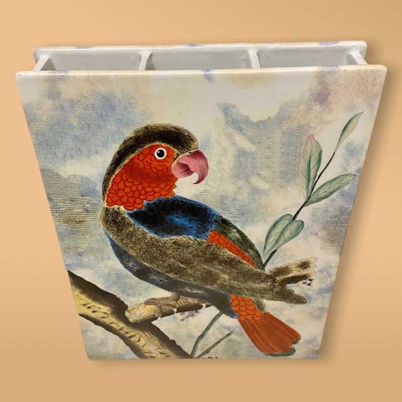 May include: A ceramic vase with a watercolor design of a parrot perched on a branch. The parrot has red, blue, and brown feathers. The vase has three compartments.
