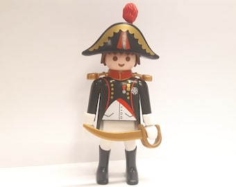May include: A plastic toy figure of a military officer wearing a black and gold uniform with a red and gold hat. The figure is holding a gold sword.