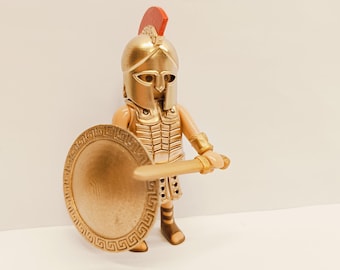 May include: A gold plastic toy soldier figurine wearing a helmet and armor, holding a sword and shield.