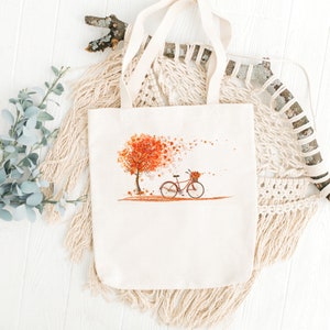 May include: A white tote bag with a design of a red bicycle with a basket of flowers and a tree with orange leaves. The leaves are blowing in the wind.