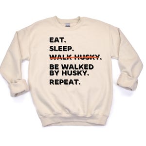 May include: A beige sweatshirt with black text that reads "Eat. Sleep. Walk Husky. Be walked by Husky. Repeat."