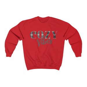 May include: A red sweatshirt with the words "COZY VIBES" printed on the front. The word "COZY" is in a plaid pattern, and "Vibes" is in a teal script font. The sweatshirt has a crew neck and long sleeves.