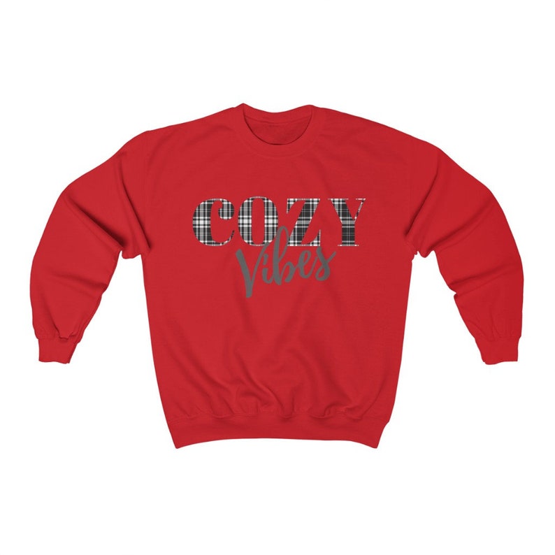 May include: A red sweatshirt with the words "COZY VIBES" printed on the front. The word "COZY" is in a plaid pattern, and "Vibes" is in a teal script font. The sweatshirt has a crew neck and long sleeves.