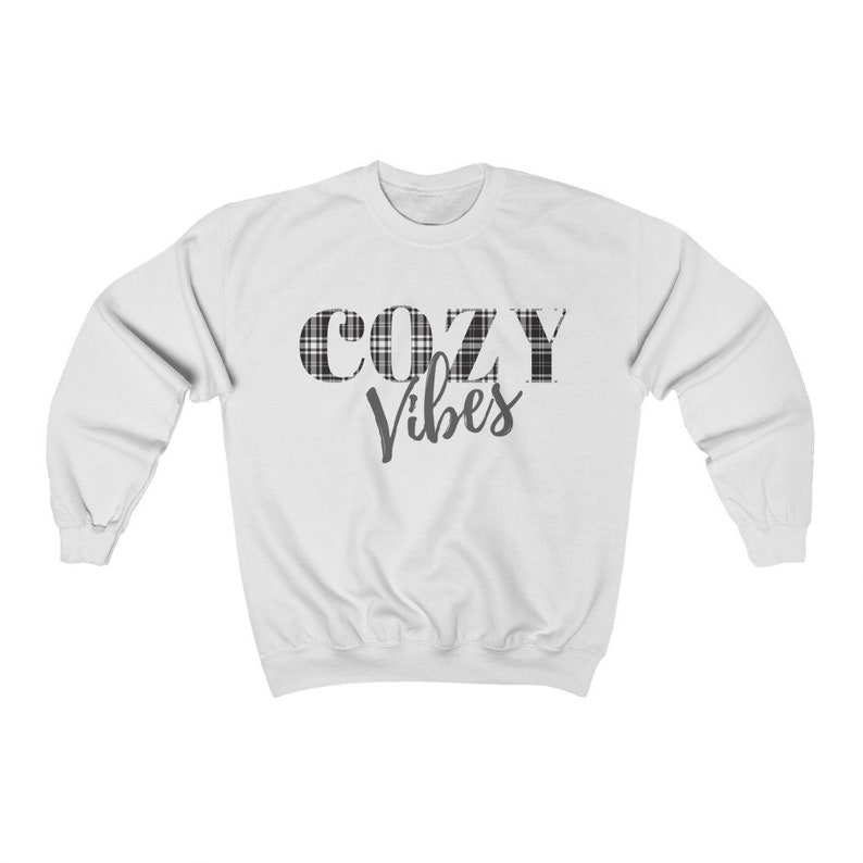 May include: White sweatshirt with the words "COZY Vibes" printed on the front. The word "COZY" is in a black and white plaid pattern. The sweatshirt has long sleeves and a crew neck.