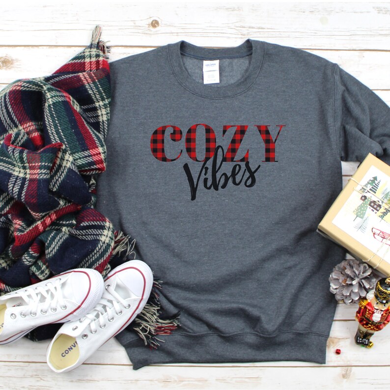 May include: A dark gray sweatshirt with the words "COZY Vibes" printed on the front. The word "COZY" is in red plaid, and "Vibes" is in black script. White sneakers and a plaid scarf are next to the sweatshirt.