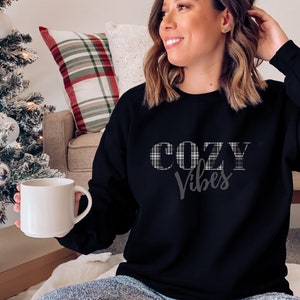 May include: Black sweatshirt with the text "COZY Vibes" in a plaid design. The model is holding a white mug. The sweatshirt is a long-sleeved top, perfect for casual wear. The background includes a Christmas tree and a plaid pillow.