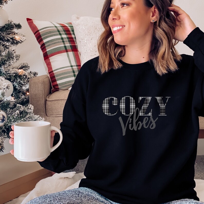 May include: Black sweatshirt with the text "COZY Vibes" in a plaid design. The model is holding a white mug. The sweatshirt is a long-sleeved top, perfect for casual wear. The background includes a Christmas tree and a plaid pillow.