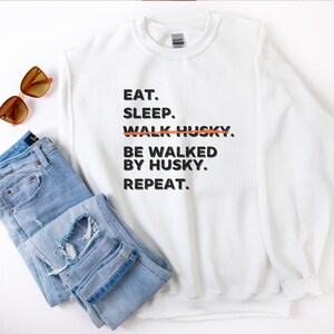 May include: White sweatshirt with black text that reads "Eat. Sleep. Walk Husky. Be walked by Husky. Repeat." The word "Walk" is crossed out with a red line.