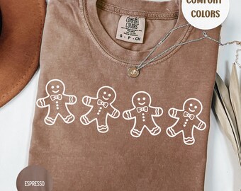 May include: Espresso-colored t-shirt with a crew neck and short sleeves. The shirt features a white graphic of four gingerbread men. The shirt has the words "Comfort Colors" printed on the tag.