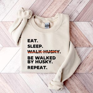 May include: A beige sweatshirt with the text "EAT. SLEEP. WALK HUSKY. BE WALKED BY HUSKY. REPEAT." printed in black. The word "WALK" is crossed out with a red line.