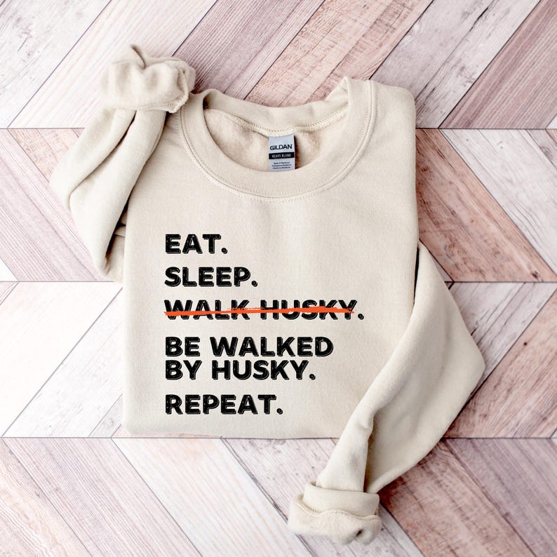 May include: A beige sweatshirt with the text "EAT. SLEEP. WALK HUSKY. BE WALKED BY HUSKY. REPEAT." printed in black. The word "WALK" is crossed out with a red line.