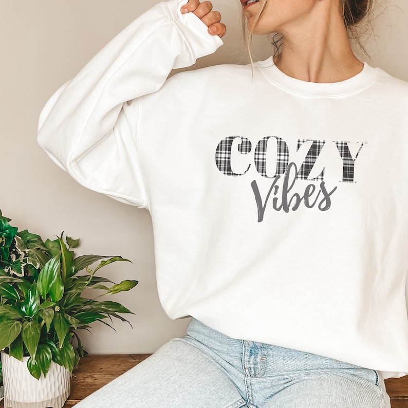 May include: White sweatshirt with the words "COZY Vibes" printed on the front. The word "COZY" is in a black and white plaid pattern. The sweatshirt is long-sleeved and the model is wearing blue jeans.