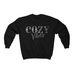 May include: Black sweatshirt with the words "COZY Vibes" printed on the front. The word "COZY" is in a plaid pattern, and "Vibes" is in a cursive font. The sweatshirt is a crew neck style.