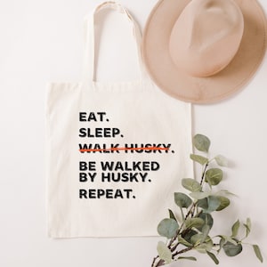 May include: Cream-colored tote bag with black text that reads "EAT. SLEEP. WALK HUSKY. BE WALKED BY HUSKY. REPEAT." The word "WALK" is crossed out in orange. A beige hat and green plant are also in the image.