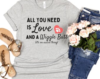 May include: A gray t-shirt with the text "ALL YOU NEED IS LOVE AND A WIGGLE BUTT (it's an aussie thing)". The design includes a red heart with a paw print. The shirt is paired with blue jeans, sandals, and a bouquet of pink roses.