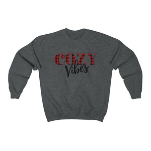 May include: Dark gray sweatshirt with the words "COZY Vibes" printed on the front. The word "COZY" is in a red and black plaid pattern, while "Vibes" is in a black script font. The sweatshirt is a crew neck style.