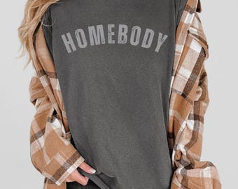 May include: A dark gray t-shirt with the word "HOMEBODY" printed in white on the front. The shirt is being worn by a person with long blonde hair and a brown and white plaid shirt.