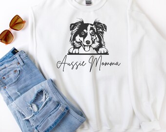May include: White sweatshirt with a black and white graphic of an Australian Shepherd dog and the text "Aussie Momma". The sweatshirt is paired with blue jeans and sunglasses.