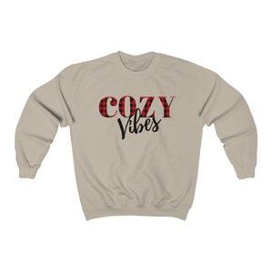 May include: A beige sweatshirt with the words "COZY Vibes" printed on the front. The word "COZY" is in red and black buffalo plaid, while "Vibes" is in a black script font. The sweatshirt has long sleeves and a crew neck.