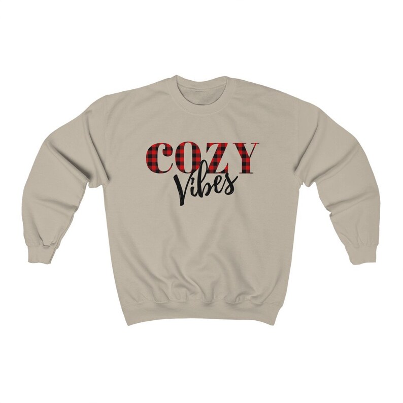 May include: A beige sweatshirt with the words "COZY Vibes" printed on the front. The word "COZY" is in red and black buffalo plaid, while "Vibes" is in a black script font. The sweatshirt has long sleeves and a crew neck.