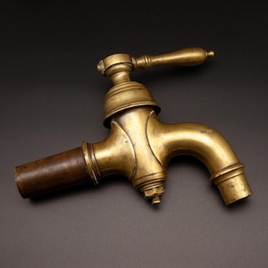 May include: Antique brass faucet with a curved spout and wooden handle. The faucet has a classic design with a cylindrical body and a lever-style handle. The finish is a warm, aged brass color, suggesting vintage appeal.