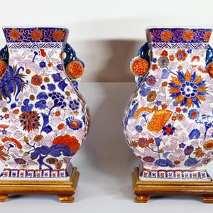 Op de afbeelding: Twee decoratieve keramische vazen met bloemmotieven in blauw, oranje en wit. Elke vaas heeft een vierkante basis en een bolvormig lichaam met handvatten. De vazen staan op een lichtgekleurd oppervlak.