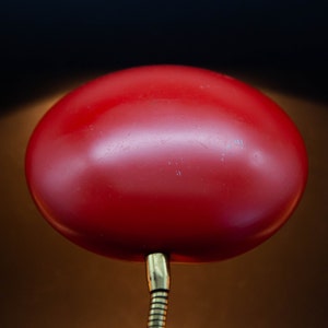 May include: A red, retro-style desk lamp with a rounded shade. The shade is a vibrant red color and is attached to a flexible, brass-colored neck. The lamp's design is reminiscent of mid-century modern aesthetics.