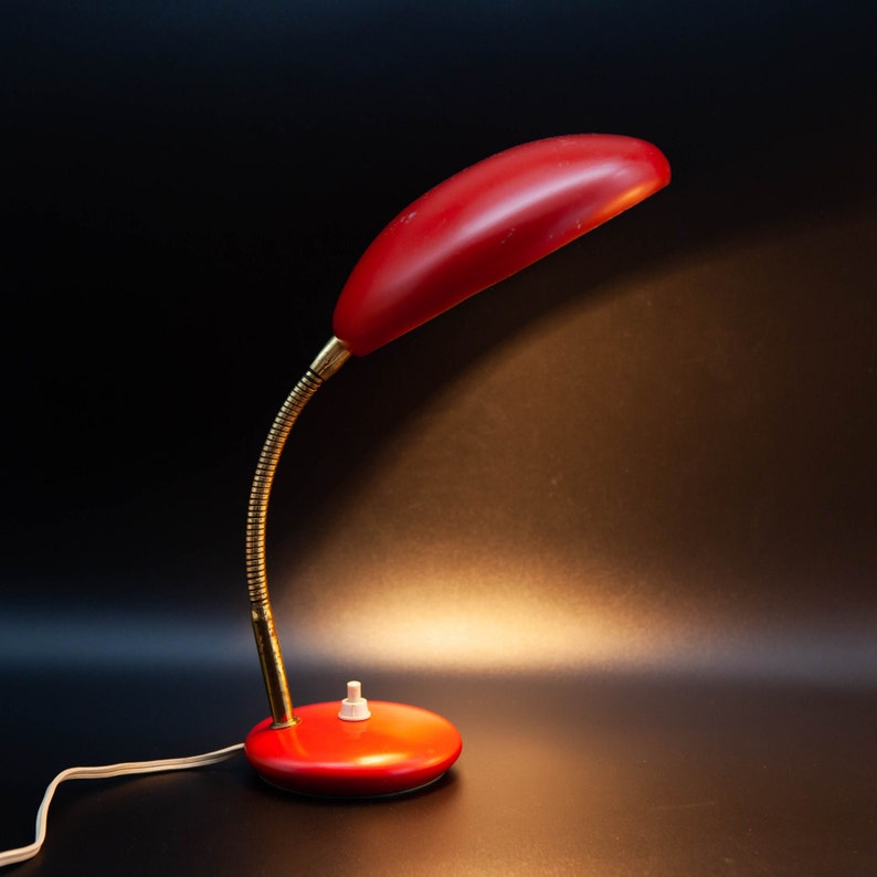 May include: A vintage red desk lamp with a flexible, brass-colored neck and a round base. The lamp has a streamlined design with a red lampshade and a white on/off switch. The lamp is plugged into a wall outlet.