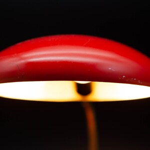 May include: A close-up of a red desk lamp with a rounded shade. The lamp is illuminated, casting a warm glow against a black background. The lamp's design is simple and modern, suitable for a desk or bedside table.