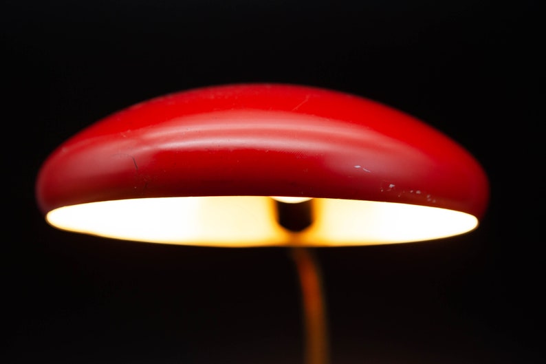 May include: A close-up of a red desk lamp with a rounded shade. The lamp is illuminated, casting a warm glow against a black background. The lamp's design is simple and modern, suitable for a desk or bedside table.