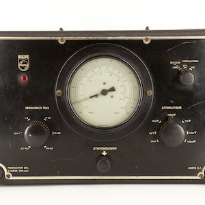 Puede incluir: Un frecuencímetro de radio Philips vintage negro con un gran dial analógico, varios botones y el texto "Frequence MHz", "Attenuateur", "Secteur Modulation", "Sortie M.F" y "SYNTONISATION".