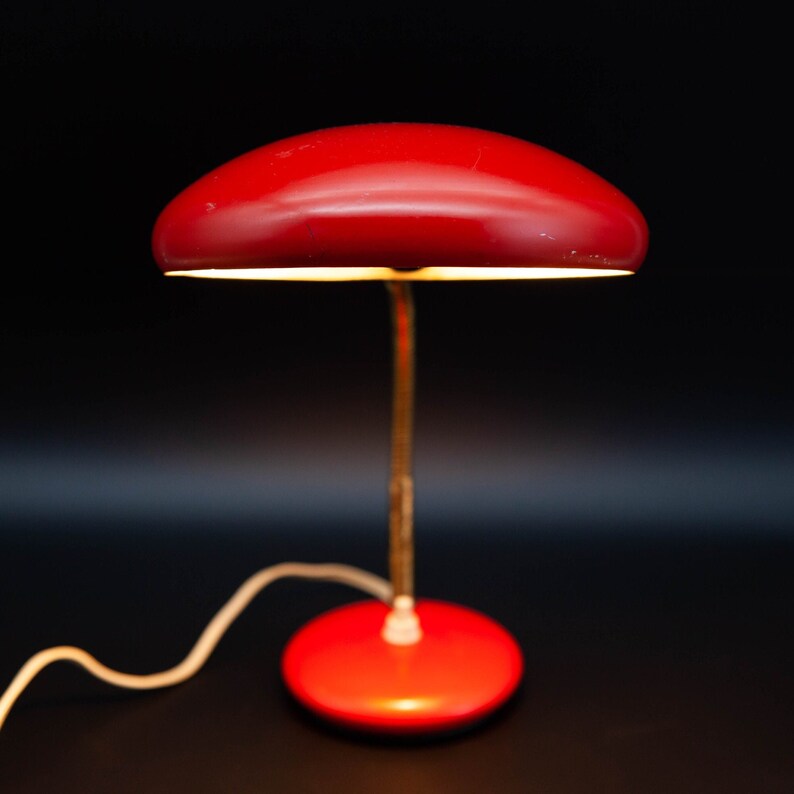May include: A vintage red desk lamp with a rounded shade and a circular base. The lamp has a slender, gold-colored stem and a white cord. The shade is positioned above the base, casting a warm glow.