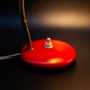 May include: Close-up of a vintage red desk lamp base with a white on/off switch. The lamp has a flexible, brass-colored neck and a white power cord. The base is a glossy, rounded shape.