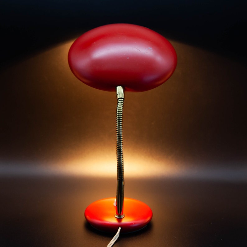 May include: A vintage red desk lamp with a flexible, gooseneck arm and a round base. The lamp has a rounded, oval-shaped shade. The lamp is illuminated, casting a warm glow against a dark background.