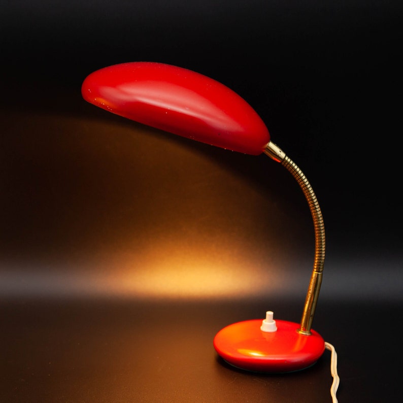 May include: A red desk lamp with a curved, oval shade and a flexible, brass-colored neck. The lamp has a round base with a white on/off switch. The cord is white. The lamp is set against a black background.