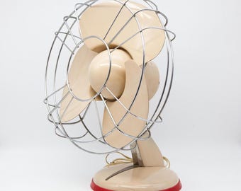 May include: Vintage beige electric fan with a chrome wire cage and red base trim. The fan has three curved blades and a rounded motor housing. A classic appliance for home decor.