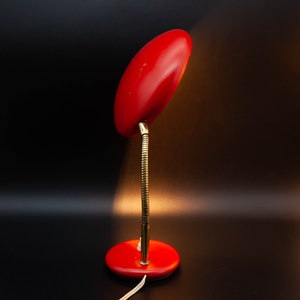 May include: A red desk lamp with a flexible, brass-colored neck and a round base. The lamp's shade is oval-shaped and also red. The lamp is plugged into a white electrical cord. The background is black.
