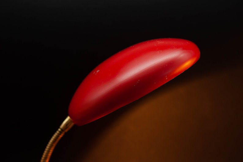 May include: A red desk lamp with a curved, elongated shade. The lamp has a flexible, brass-colored neck and is set against a dark background. The lamp's shade is illuminated, suggesting it is turned on.