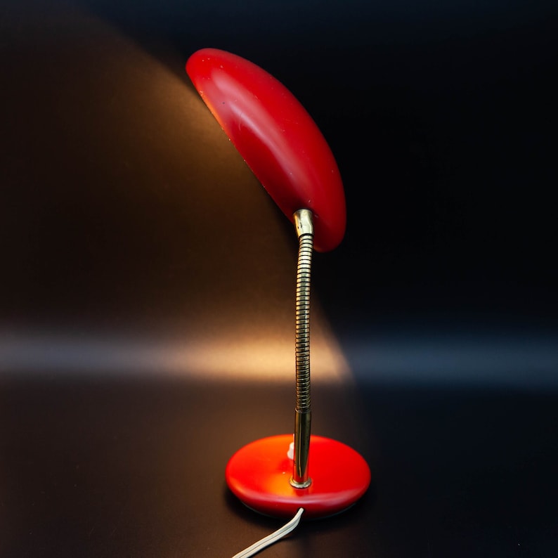 May include: A vintage red desk lamp with a flexible, brass-colored neck and a round base. The lamp's shade is oval-shaped, and the cord is white. The lamp is set against a dark background, with a spotlight effect.