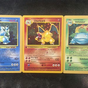 May include: Three collectible trading cards featuring Blastoise, Charizard, and Venusaur. The blue Blastoise card, red Charizard card, and green Venusaur card each display the character illustration, name, HP, and game statistics in English text.