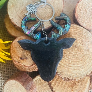 May include: A black deer head keychain with iridescent abalone shell antlers. The keychain has a silver clasp and a small tree charm. The deer head is made of resin and is displayed on a wooden background.