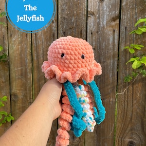 May include: A crocheted jellyfish toy with a peach-colored body and teal tentacles. The jellyfish is being held by a hand in front of a wooden fence.