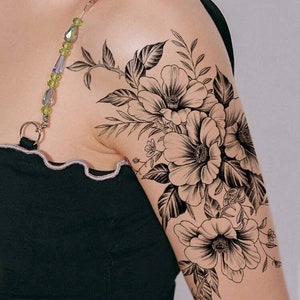 May include: Black and white floral tattoo design featuring three large flowers with detailed petals and leaves. The tattoo is on a person's upper arm.